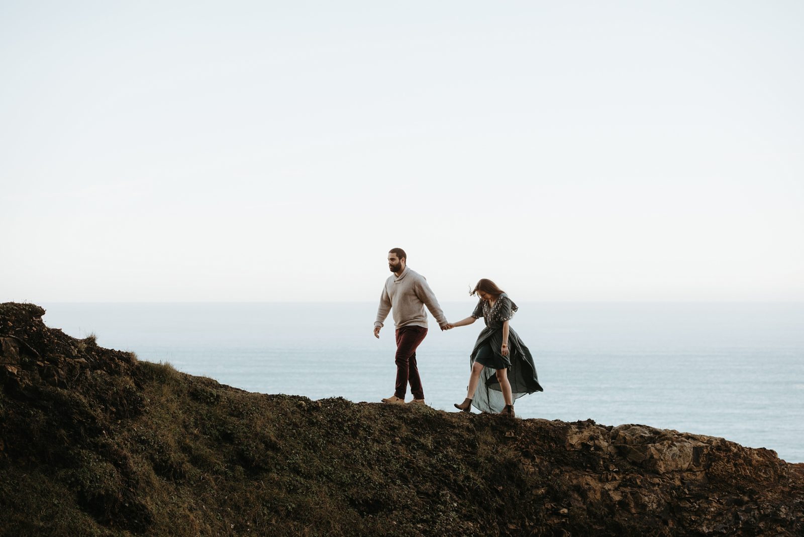 Couple walking along cliff side in flowy dress during engagement session on the Oregon Coast
