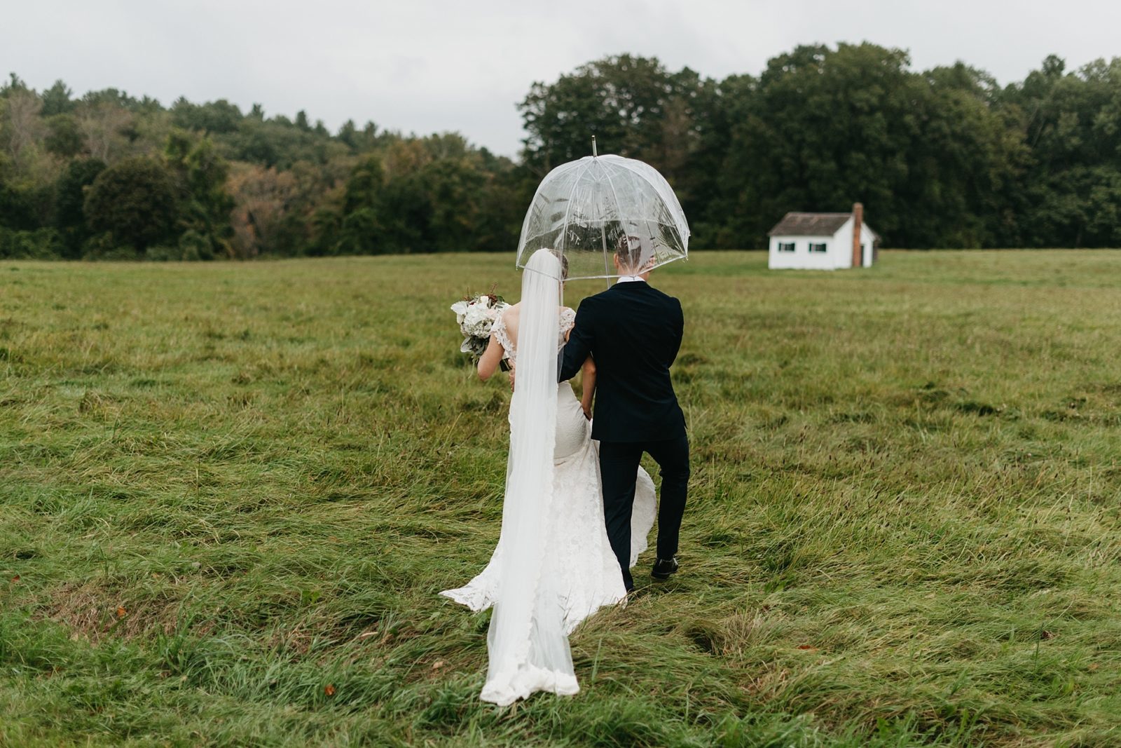 Classy couple walking through grassy field with umbrella towards a small white house in New England