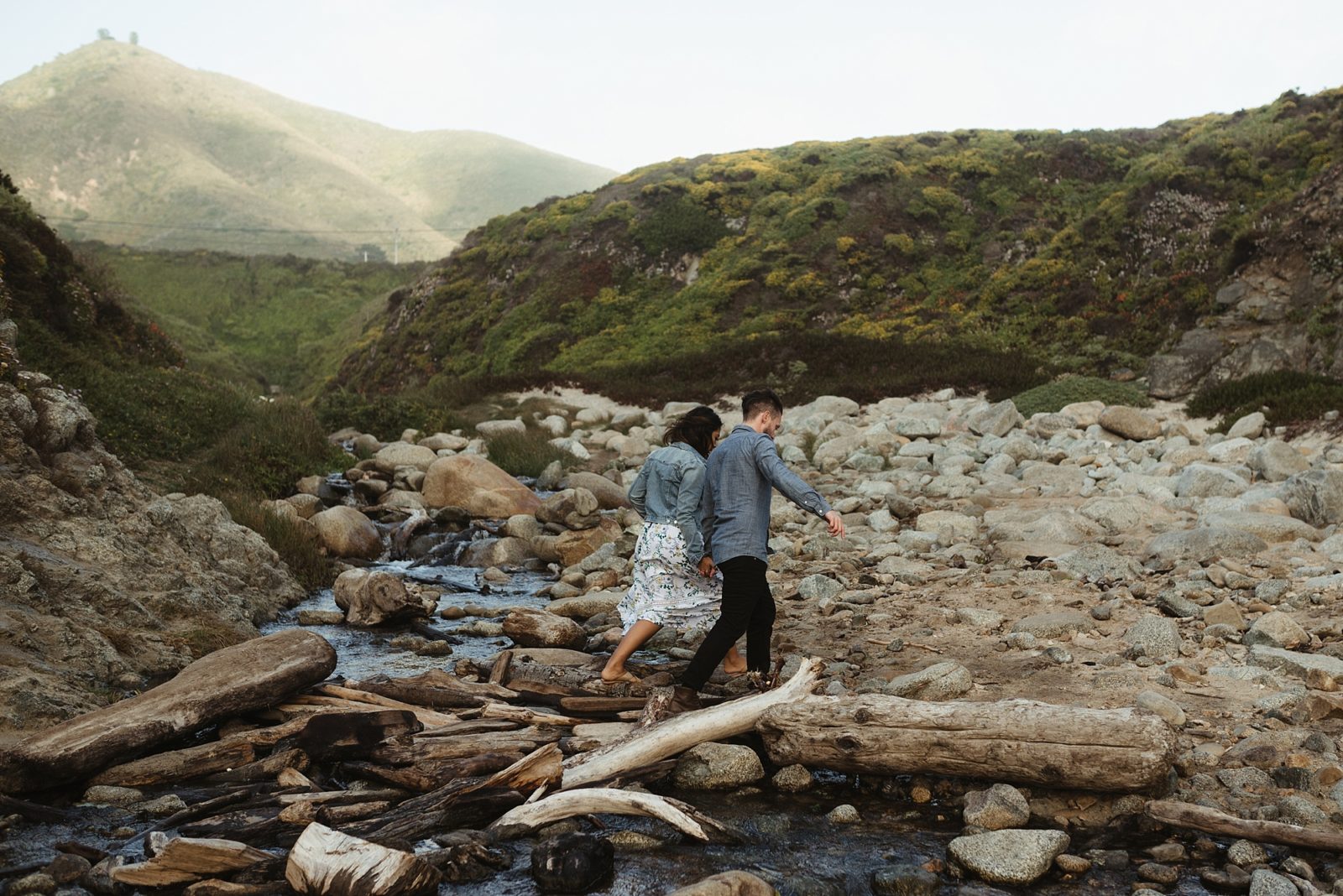 Couple walking on driftwood on the beach in Big Sur with green hills in the background during engagement shoot